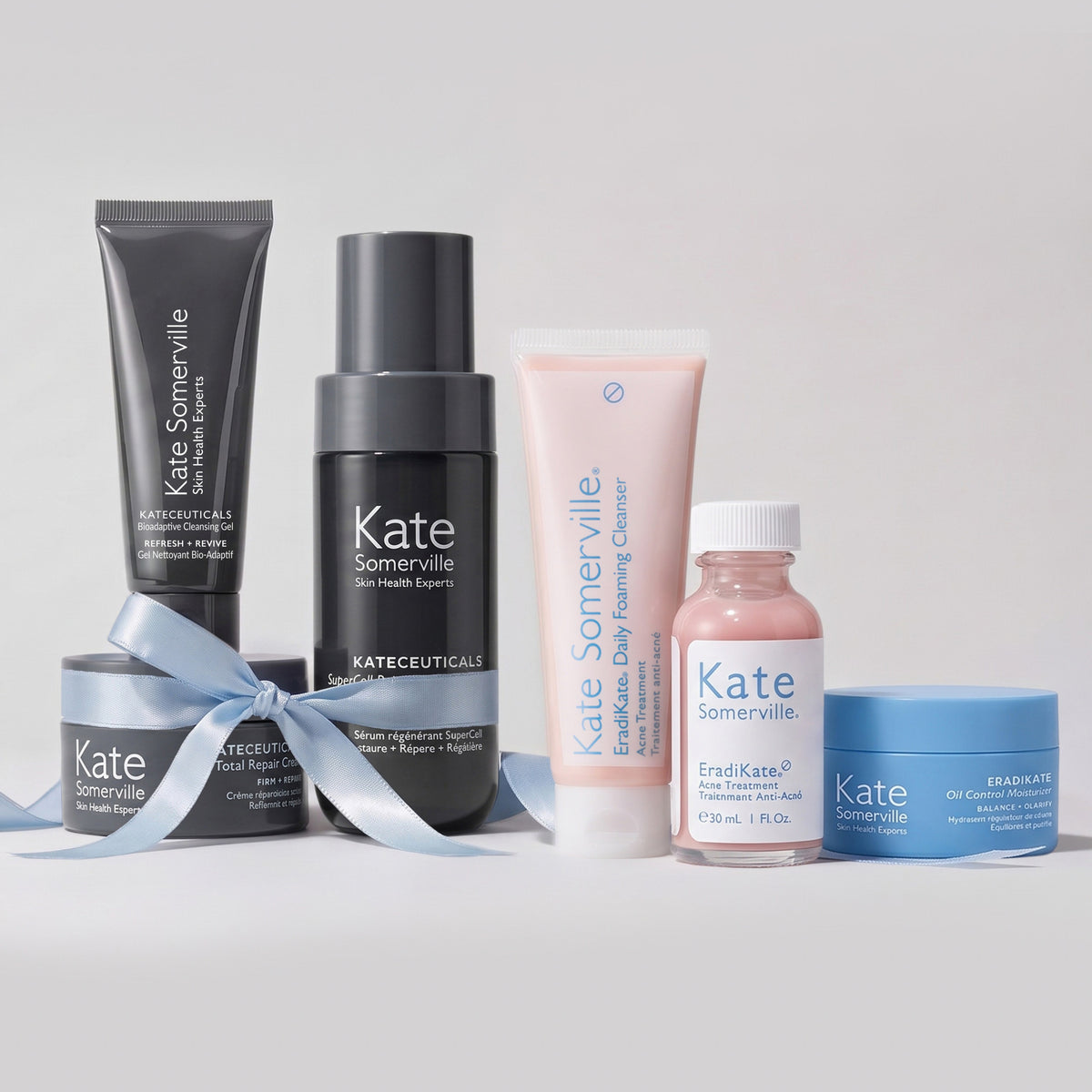 Kate Somerville holiday gift sets: the Lifted + Luminous Set with Bioadaptive Cleansing Gel, SuperCell serum and Total Repair Cream tied with a blue ribbon, beside the Clear Skin Set with EradiKate Daily Foaming Cleanser, Acne Treatment and Oil Control Moisturizer.
