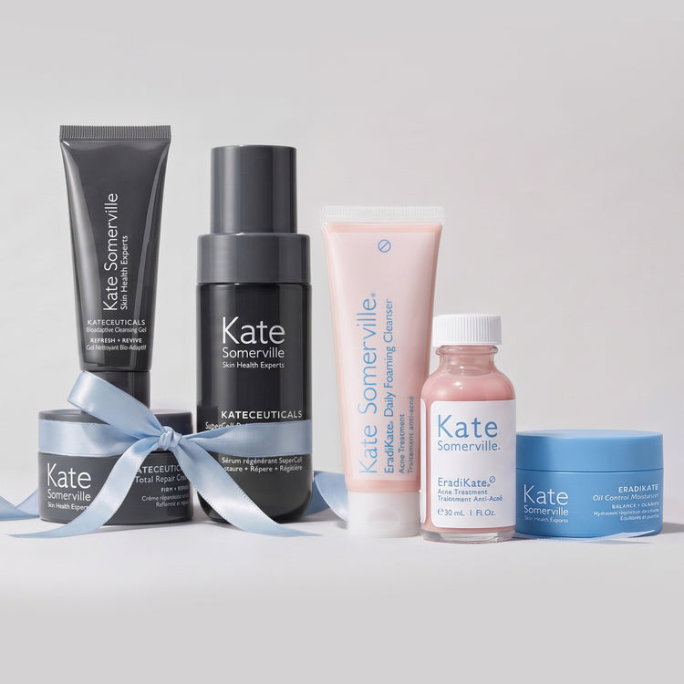 Kate Somerville holiday gift sets: the Lifted + Luminous Set with Bioadaptive Cleansing Gel, SuperCell serum and Total Repair Cream tied with a blue ribbon, beside the Clear Skin Set with EradiKate Daily Foaming Cleanser, Acne Treatment and Oil Control Moisturizer.