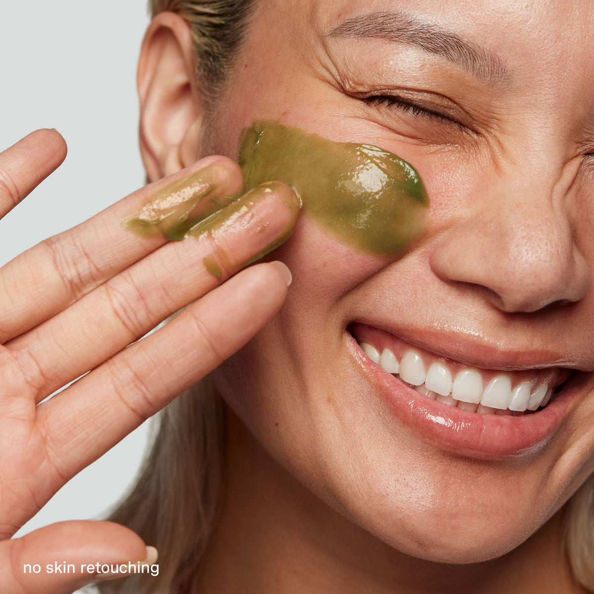 Smiling model applying green ExfoliKate Intensive scrub to her cheek with her fingertips